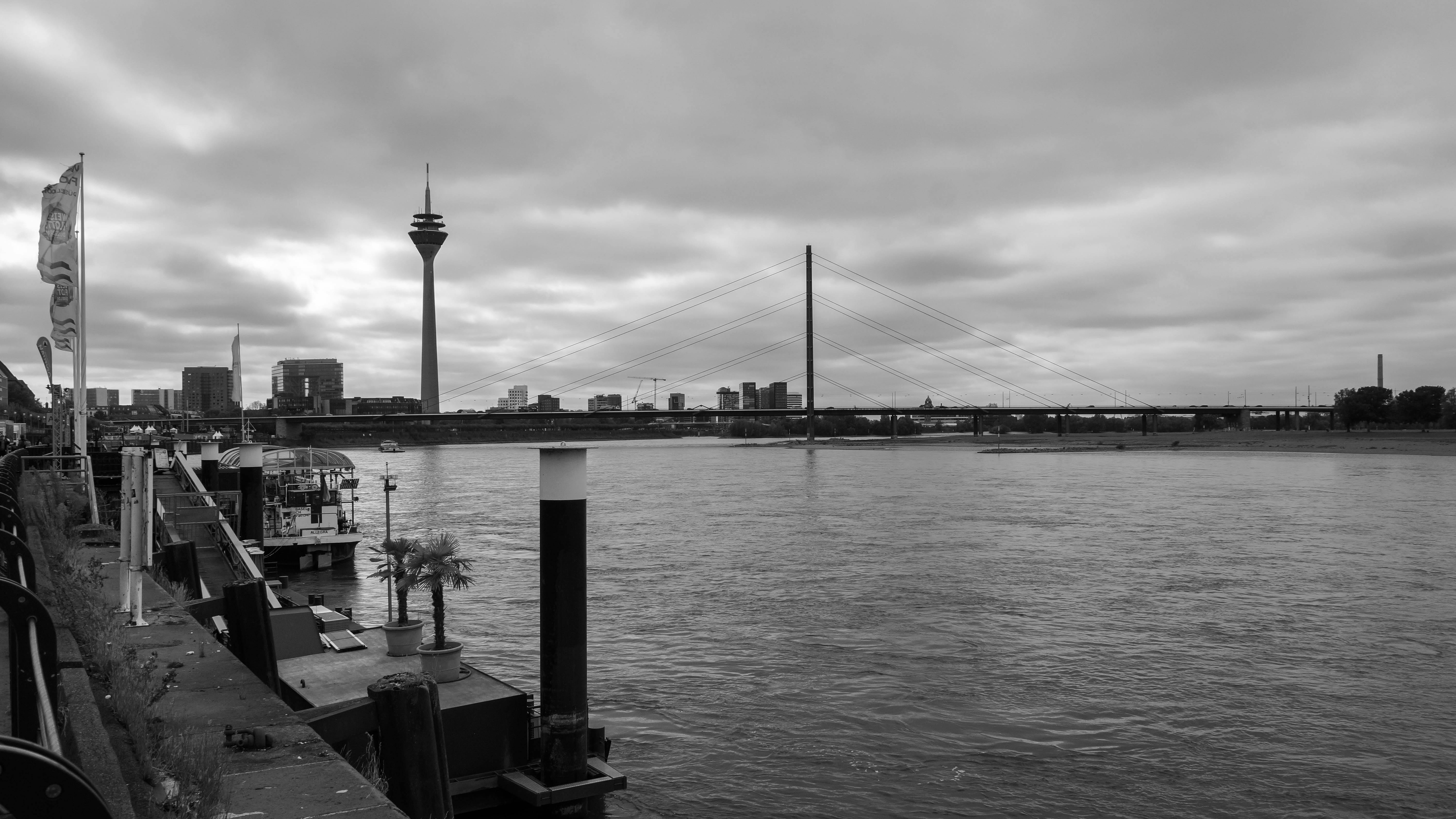 Black and white view of Düsseldorf's Rhine River with Rheinturm and bridge on a cloudy day.