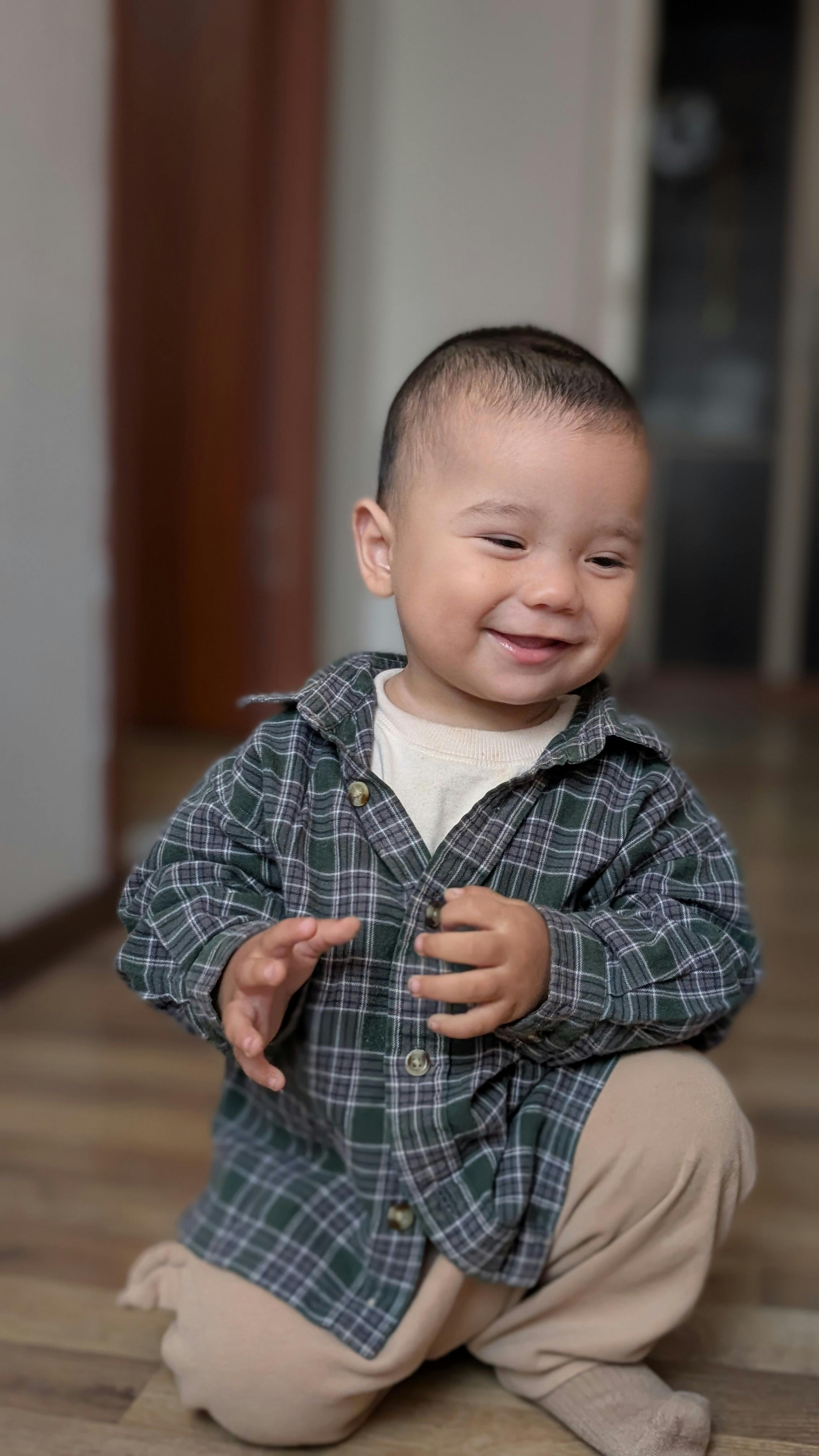 Adorable baby wearing plaid shirt smiling warmly indoors on wooden floor.