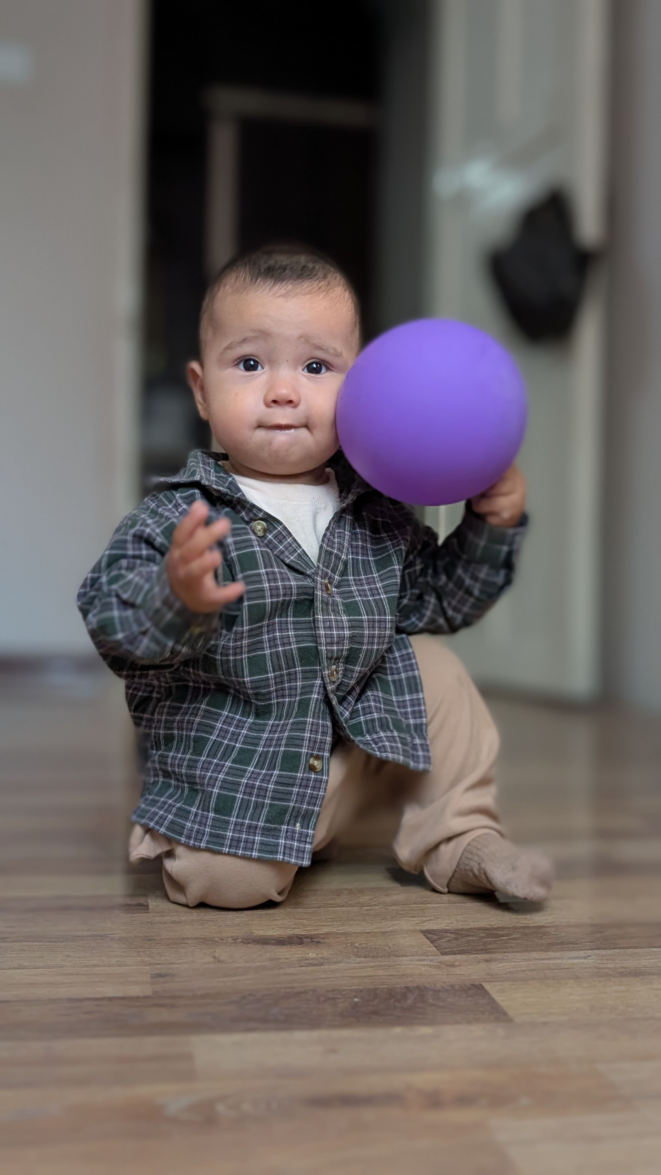 Cute baby dressed in plaid, playing with a purple balloon on wooden floor indoors.