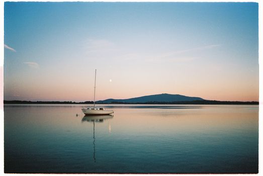 Peaceful scene of a sailboat on a calm lake with a stunning sunset backdrop.