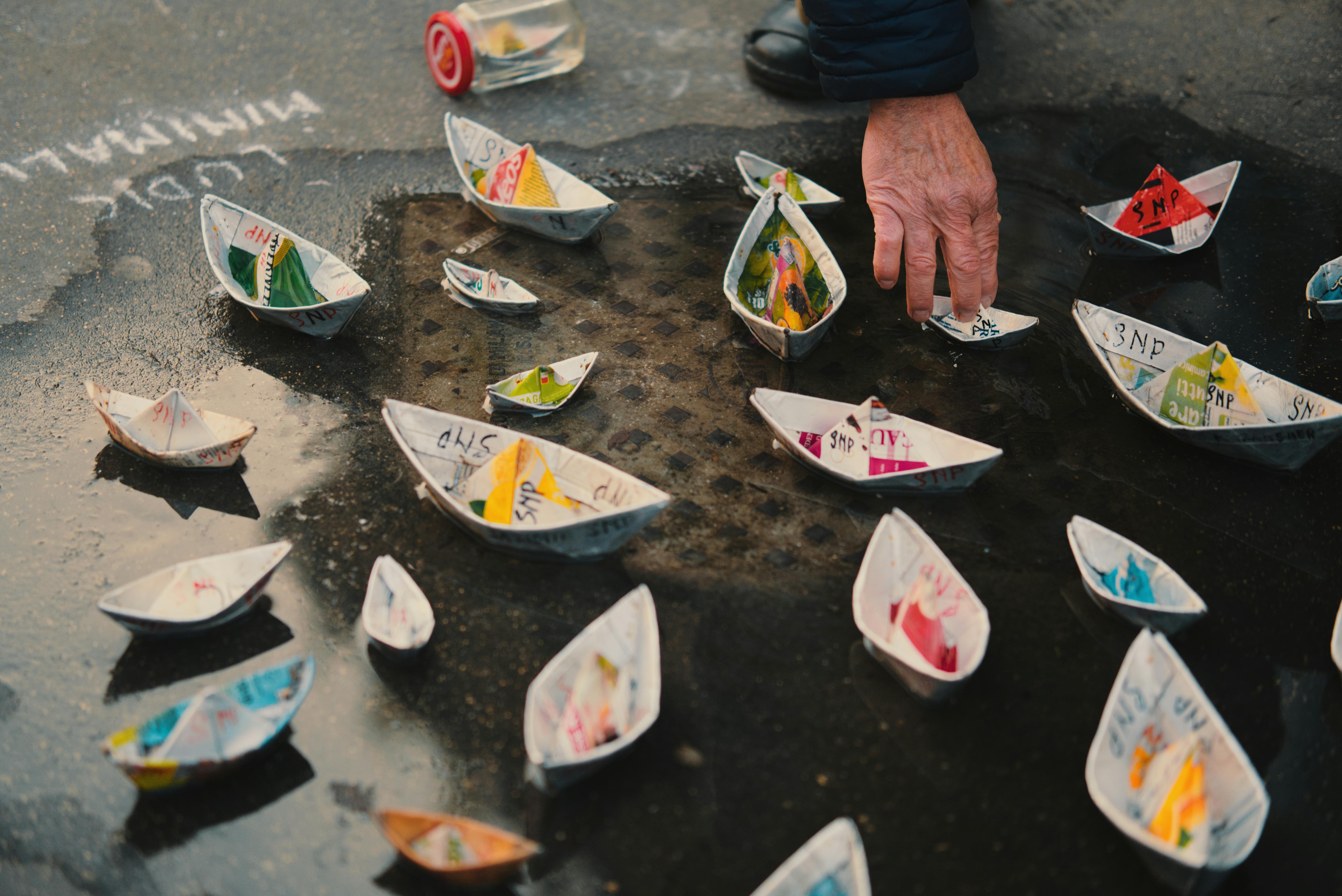 A hand arranges colorful paper boats on a wet street in Milan, Italy, capturing a whimsical moment.