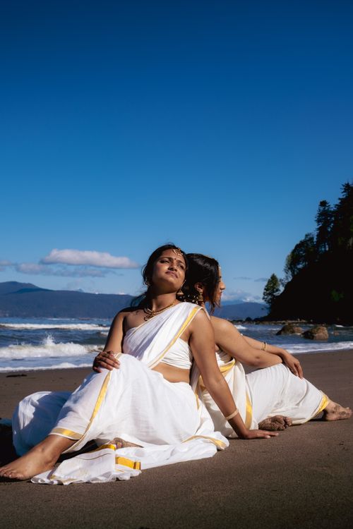 Free Two South Asian women in traditional sarees sit on Vancouver beach, embracing cultural fashion by the ocean. Stock Photo