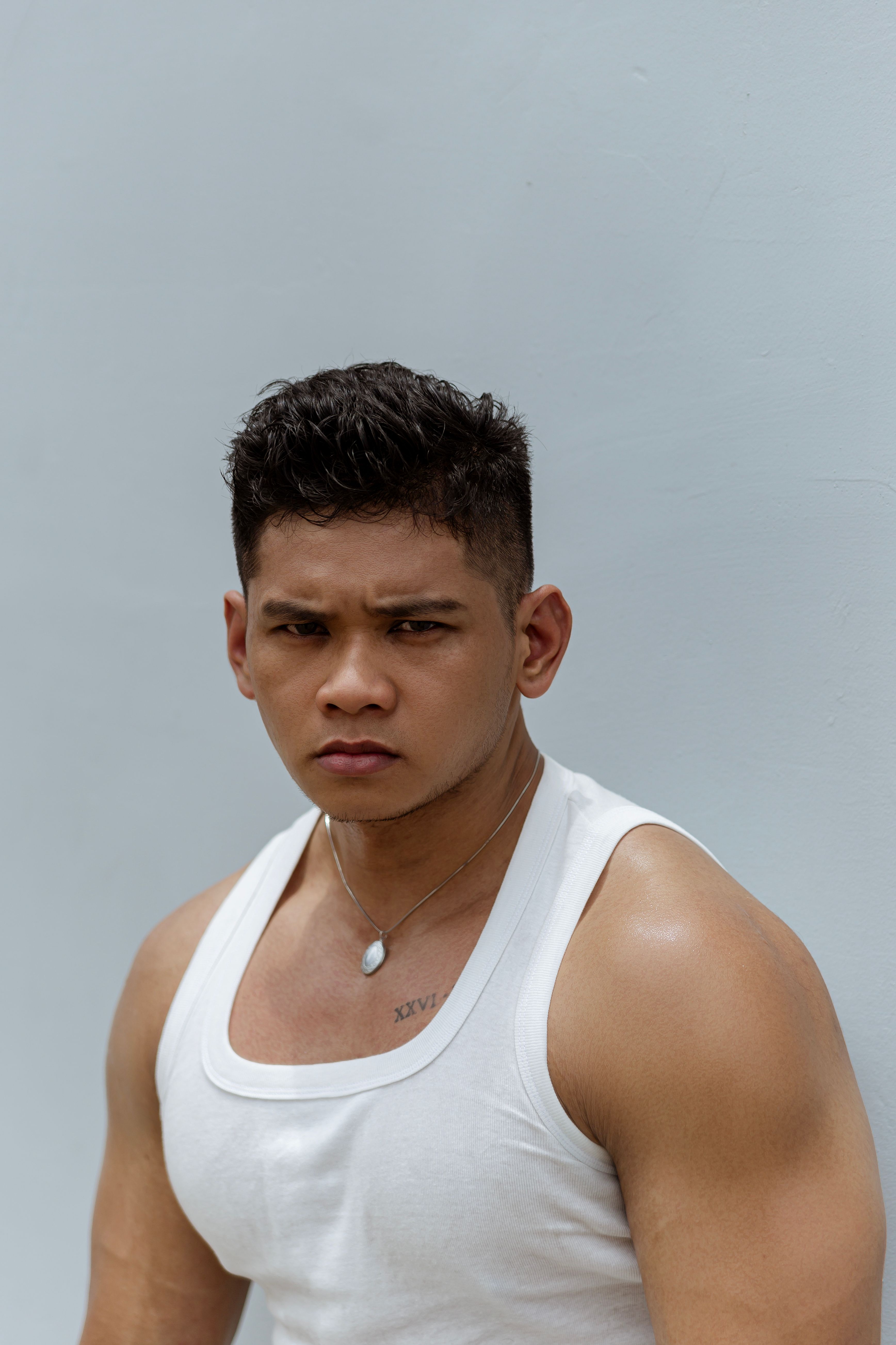 A strong male poses in a white tank top against a plain background, exuding confidence and strength.