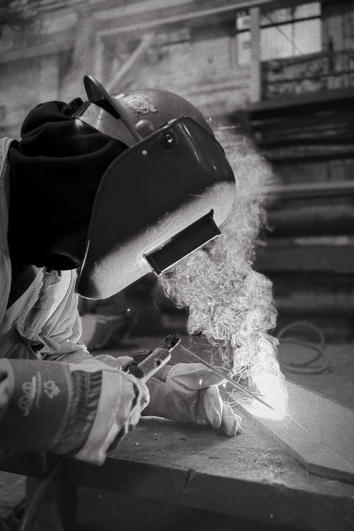 Free Black and white image of a welder creating sparks, wearing protective gear in an industrial setting. Stock Photo
