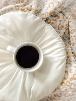 A steaming cup of coffee atop fluffy pillows and a leopard print blanket, exuding warmth and relaxation.