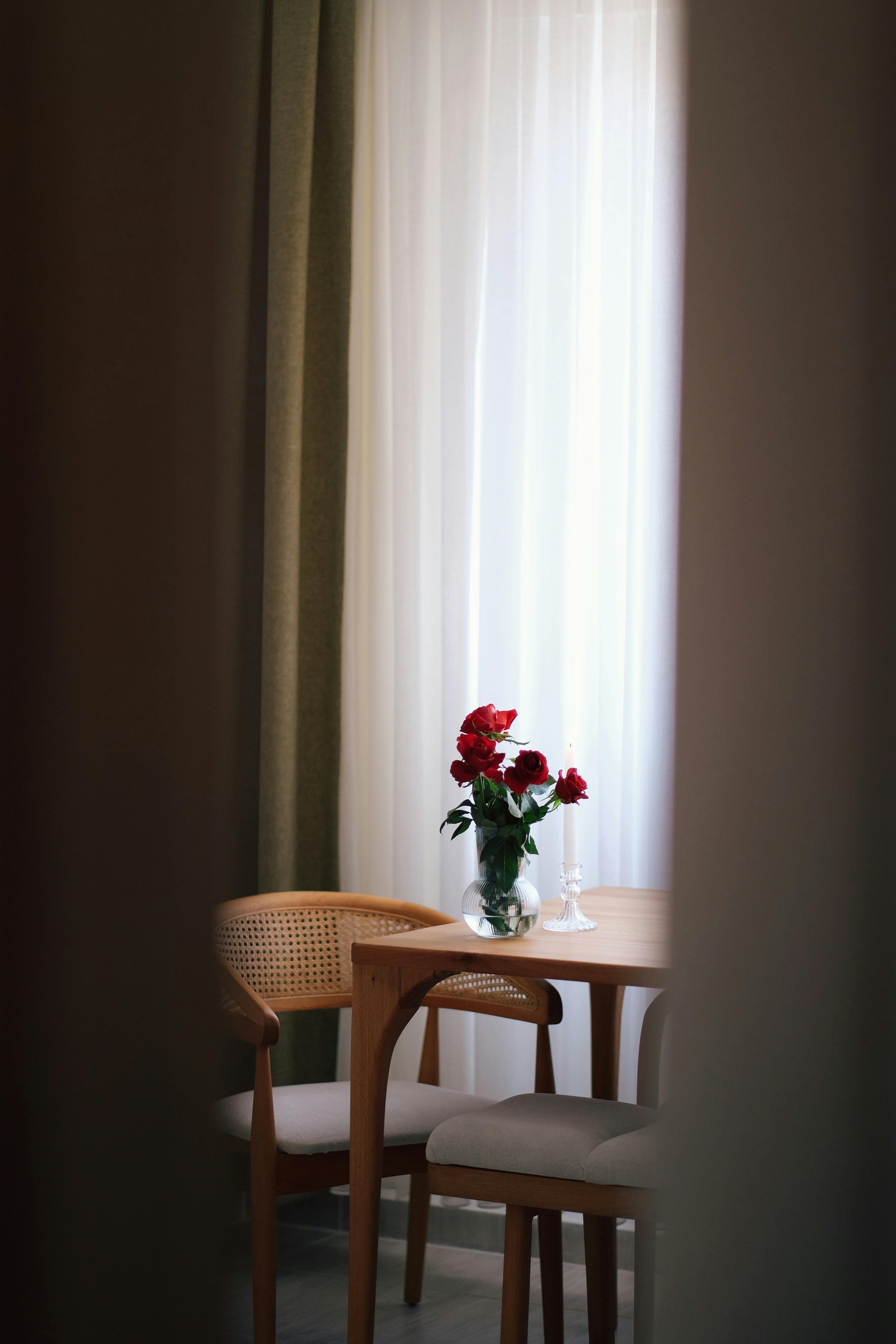 A cozy indoor setting featuring a wooden table with red roses in a glass vase by a sunlit window.