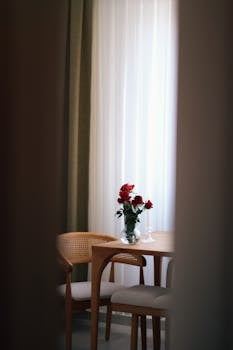 A cozy indoor setting featuring a wooden table with red roses in a glass vase by a sunlit window.