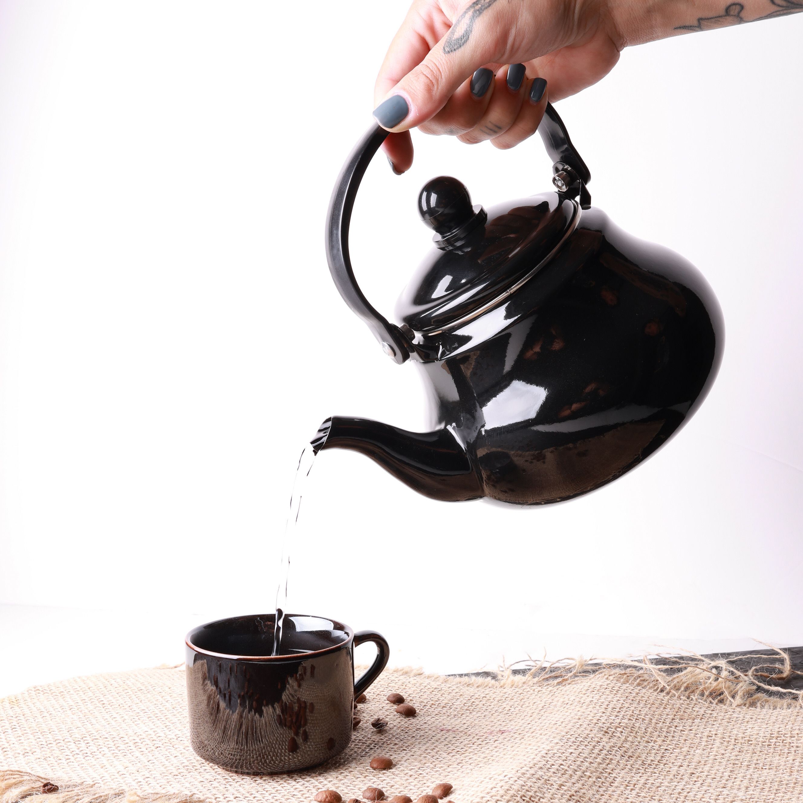 Hand pouring beverage from black teapot into ceramic mug on textured mat.