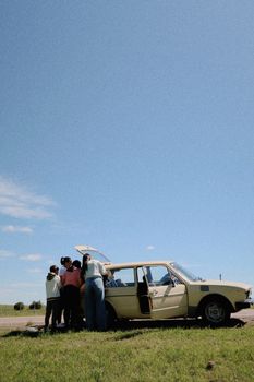 A group enjoying a road trip by a vintage car on a sunny day in Melo, Uruguay.