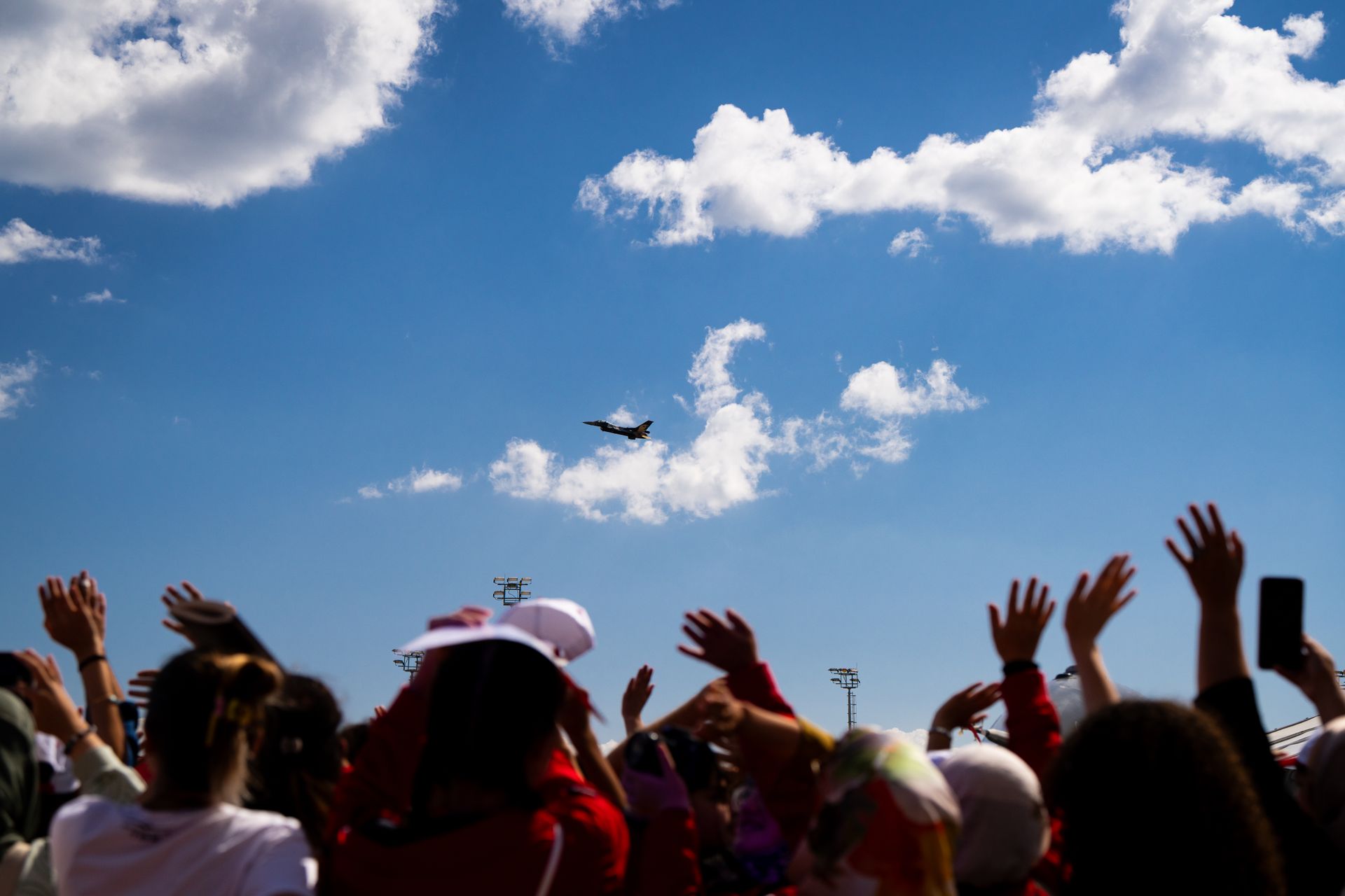 A vibrant crowd raises hands in excitement as a fighter jet performs under a clear blue sky at an airshow.