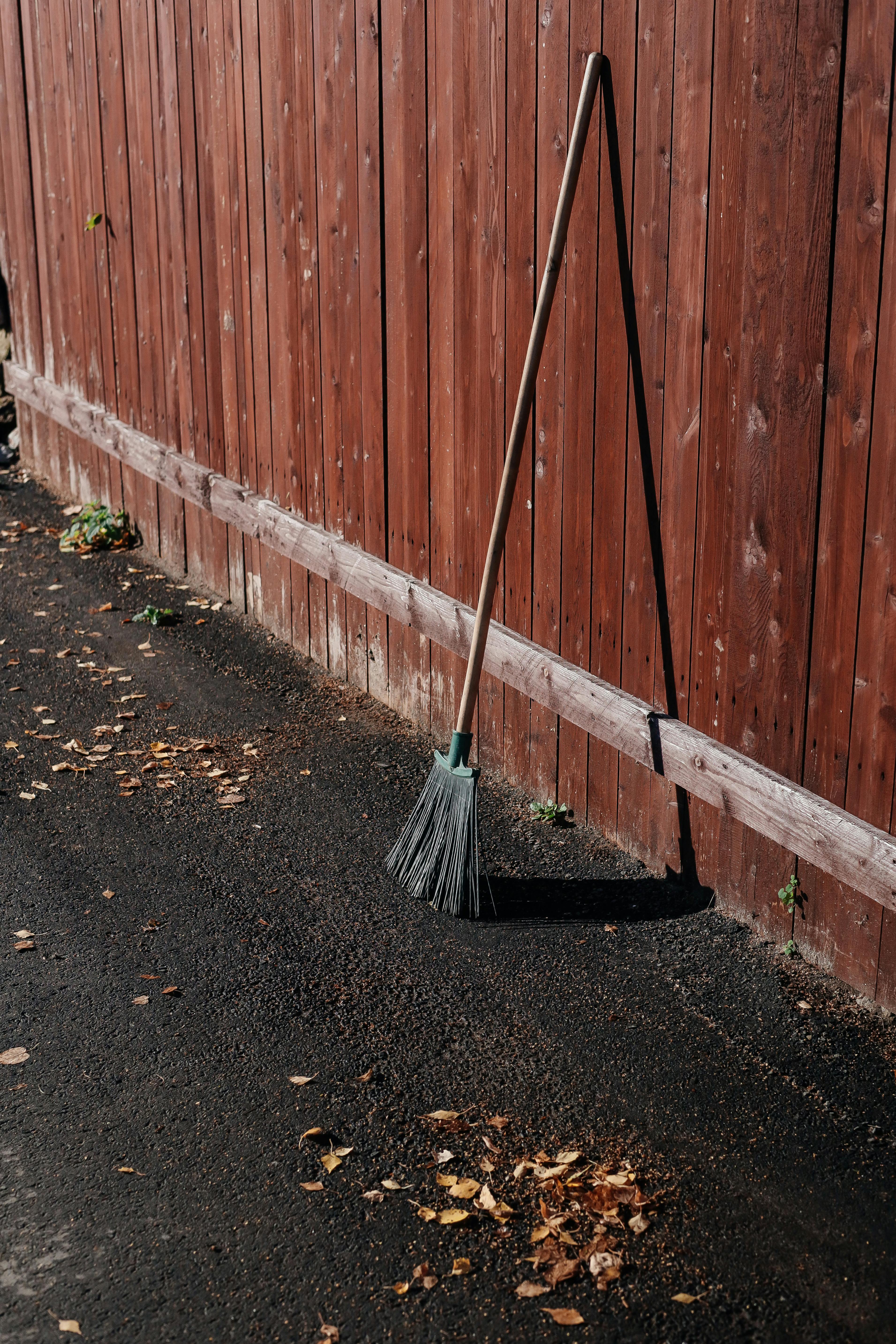 A solitary broom leaning against a red wooden fence on a sunny autumn day.