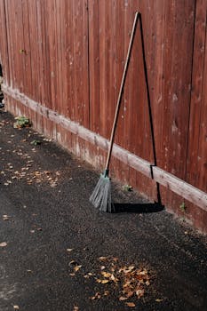 A solitary broom leaning against a red wooden fence on a sunny autumn day.