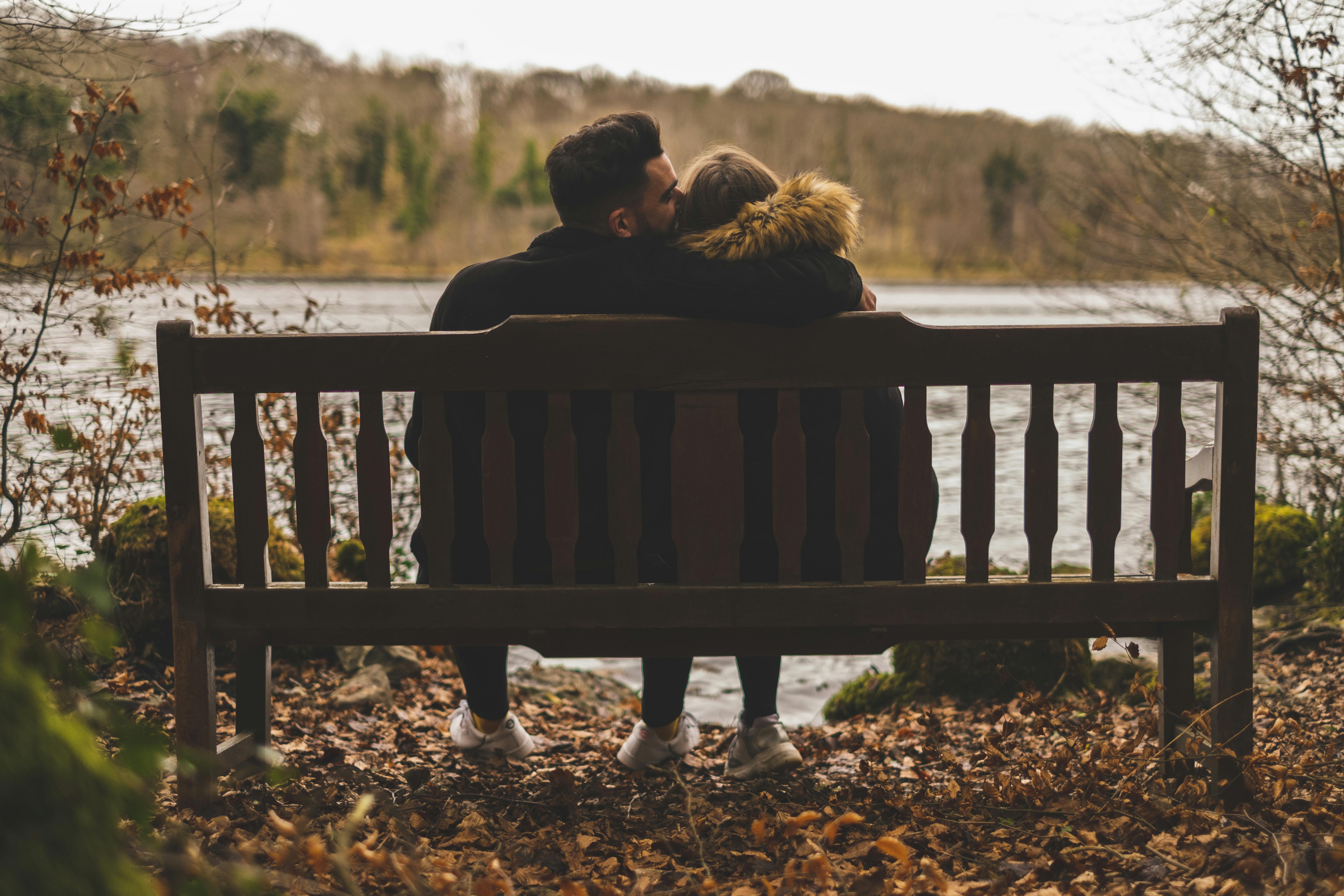 man kissing woman s head while sitting on bench in front of body of water