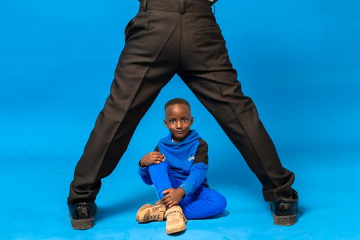 Young boy in blue tracksuit sits confidently between adult's legs against a blue backdrop in studio.