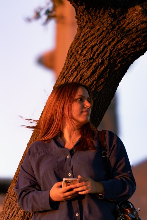 Free A thoughtful woman leans against a tree in Assos, basking in the evening light, holding her phone. Stock Photo