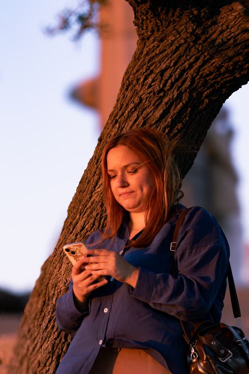 Free Young woman using smartphone leaning against tree during sunset in Assos, Türkiye. Stock Photo