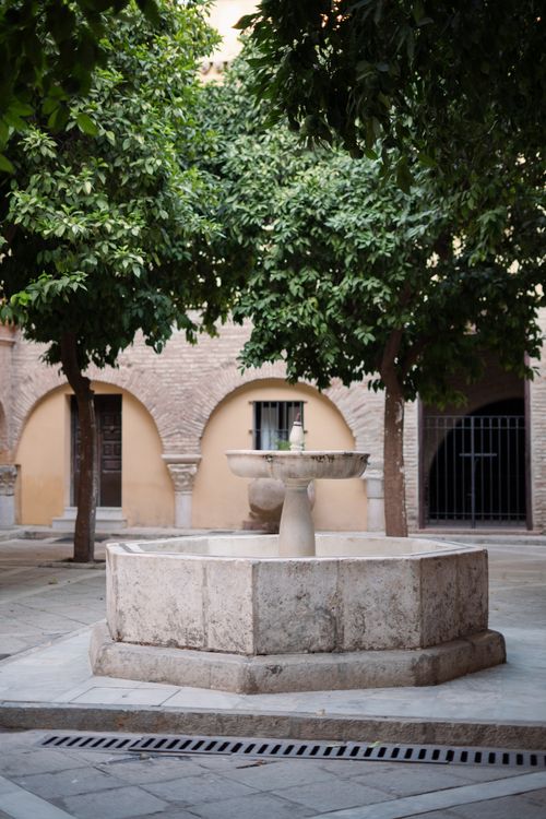 Free A peaceful courtyard with a stone fountain surrounded by lush trees and arches. Stock Photo