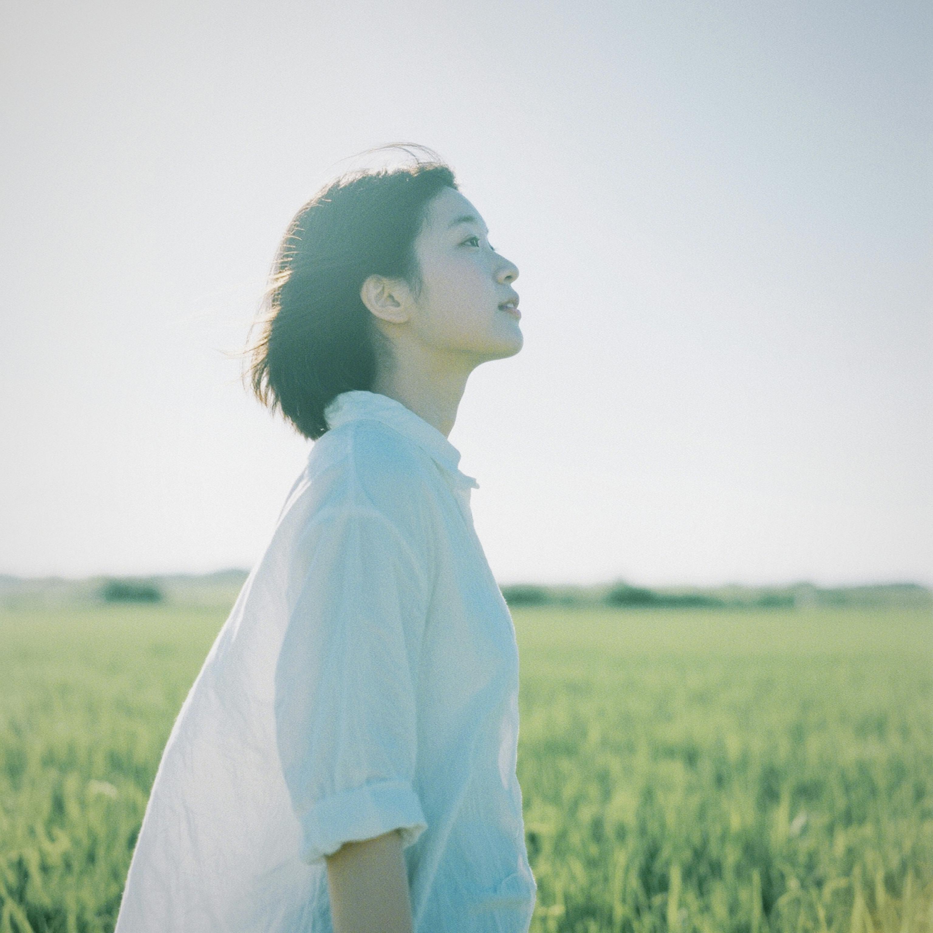 A woman in a white shirt gazes peacefully in a lush green field on a sunny day.