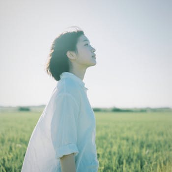 A woman in a white shirt gazes peacefully in a lush green field on a sunny day.