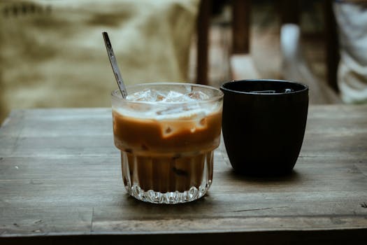 Glass of iced coffee with a spoon beside a black cup on rustic wooden table.