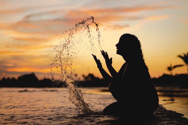 Silhouette Of Woman Sitting On The Beach Splashing Water