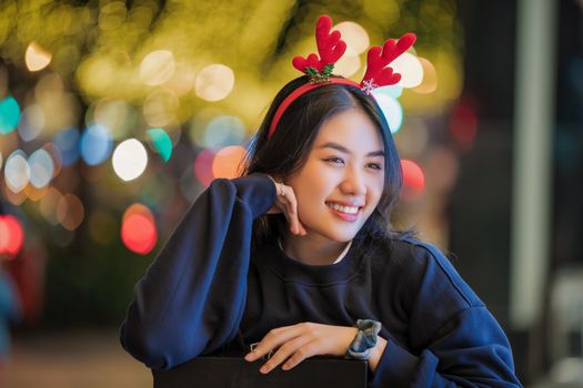 Young woman with reindeer antlers headband, smiling against festive bokeh lights at night.