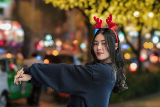 Young woman wearing red reindeer antlers, smiling at night with city lights background.