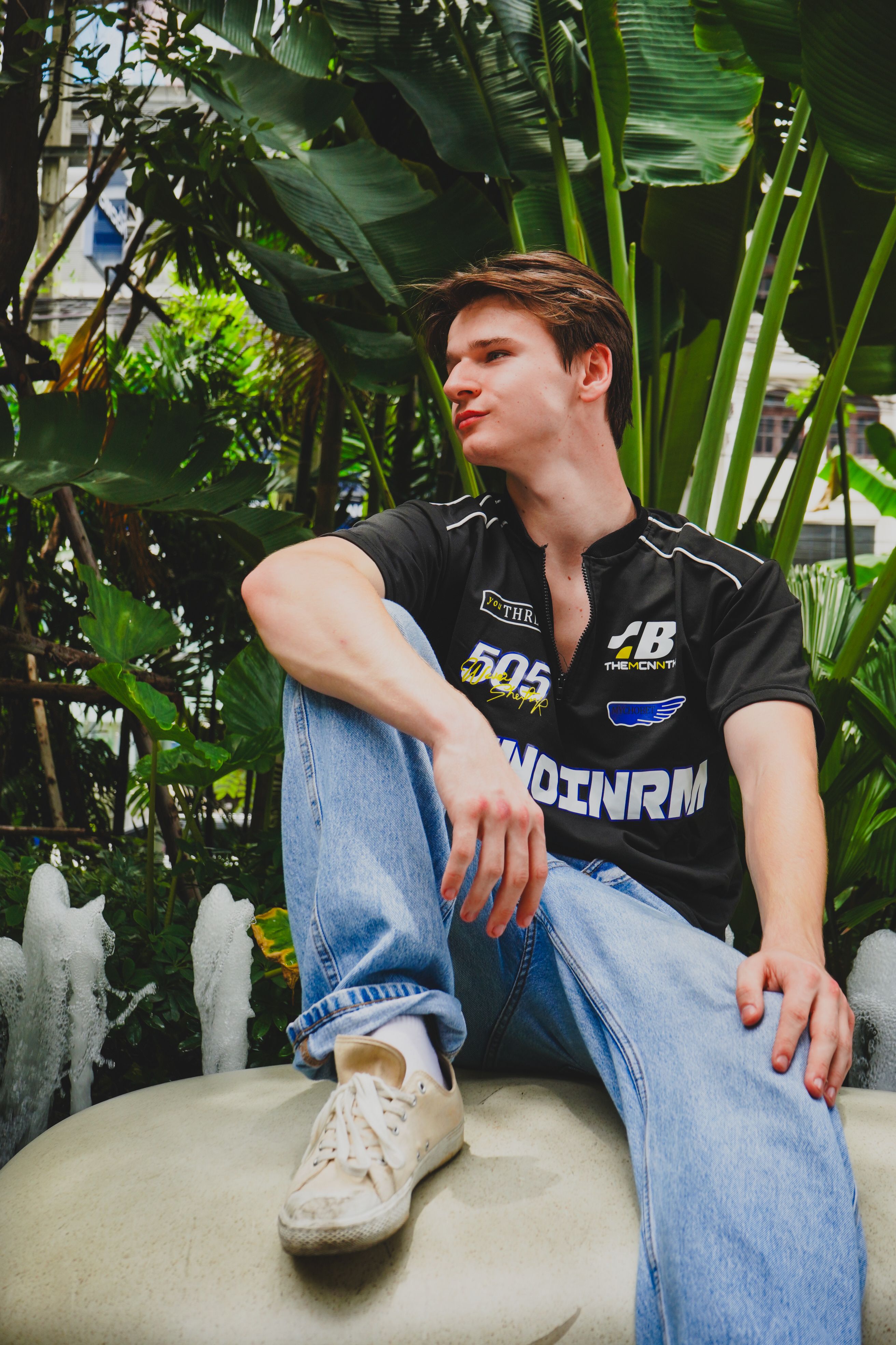 Young man in casual attire sitting among lush tropical plants outdoors.