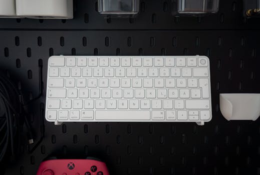 Top view of a white wireless keyboard against a perforated black surface, with red controller.