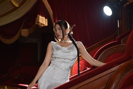 Elegant woman posing in a luxurious theater wearing a white dress, exuding sophistication.