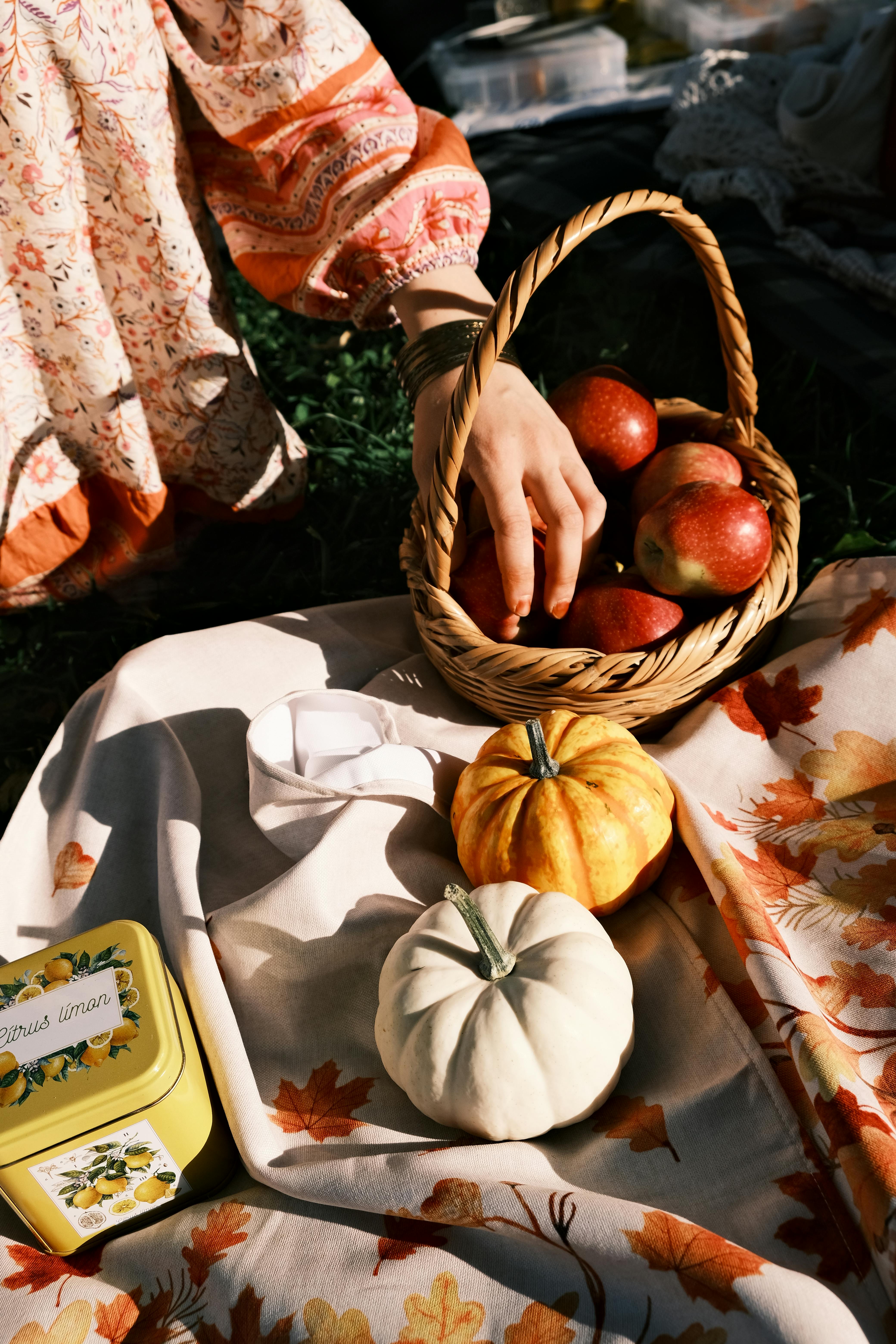 Cozy autumn picnic scene with apples in a basket and small pumpkins on a leaf-patterned cloth.