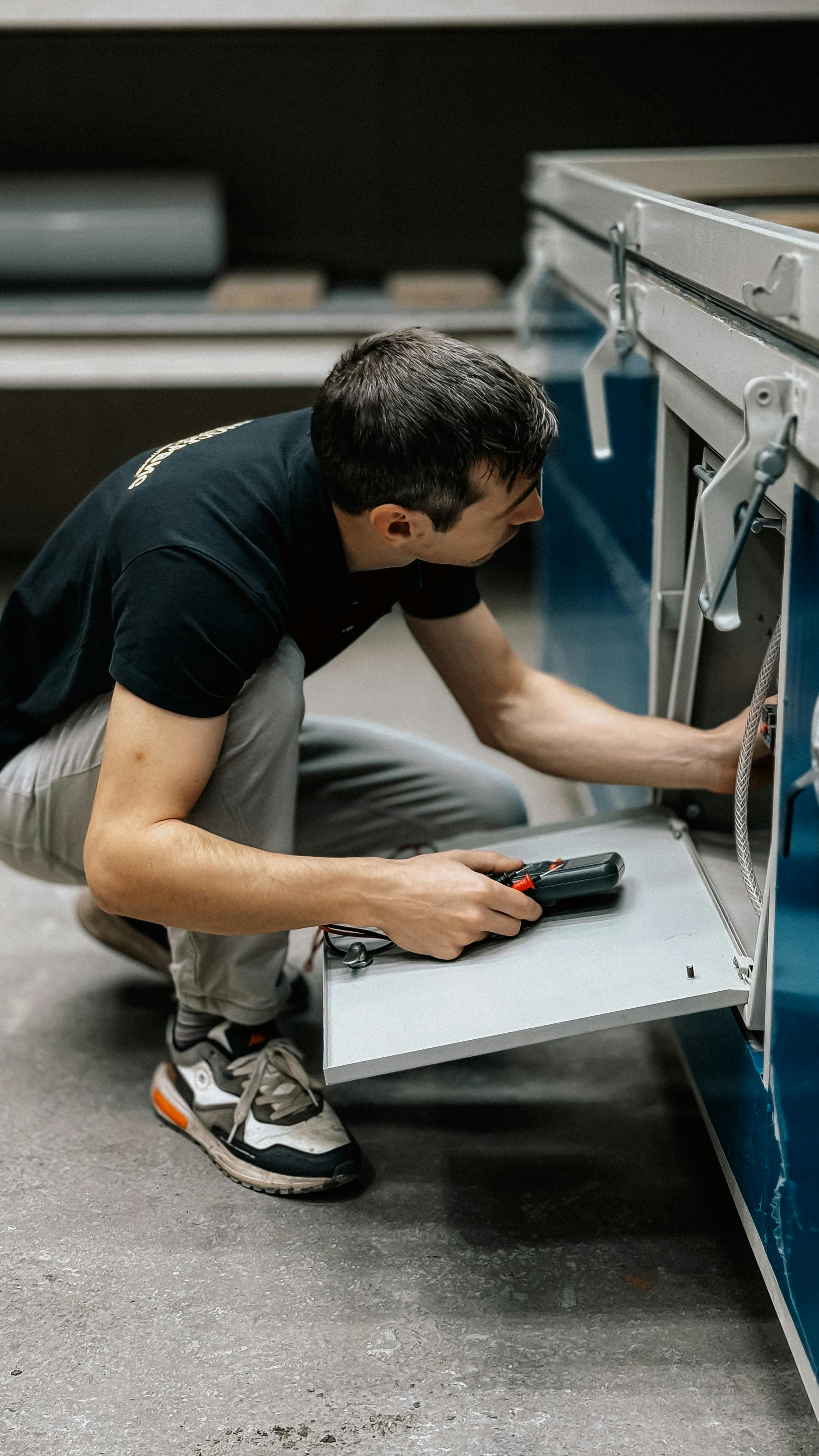 Man in black shirt and gray pants crouches by a machine, holding a device, appearing to work.