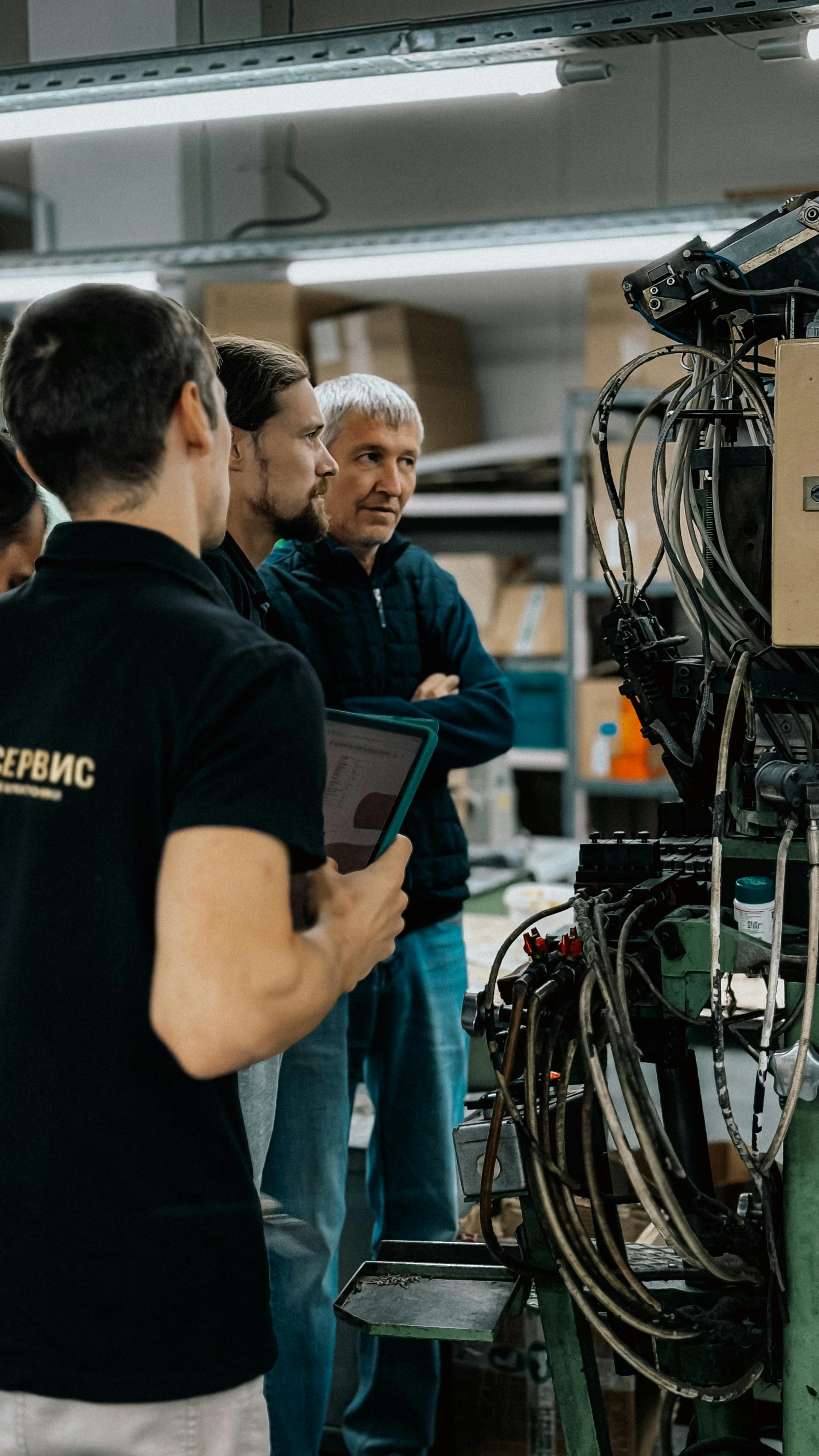 Group of people observing machinery, one holding a tablet, in a workshop setting.