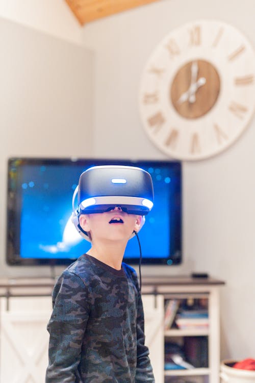 Curious little boy in VR goggles and casual clothes looking up while experiencing virtual reality at home