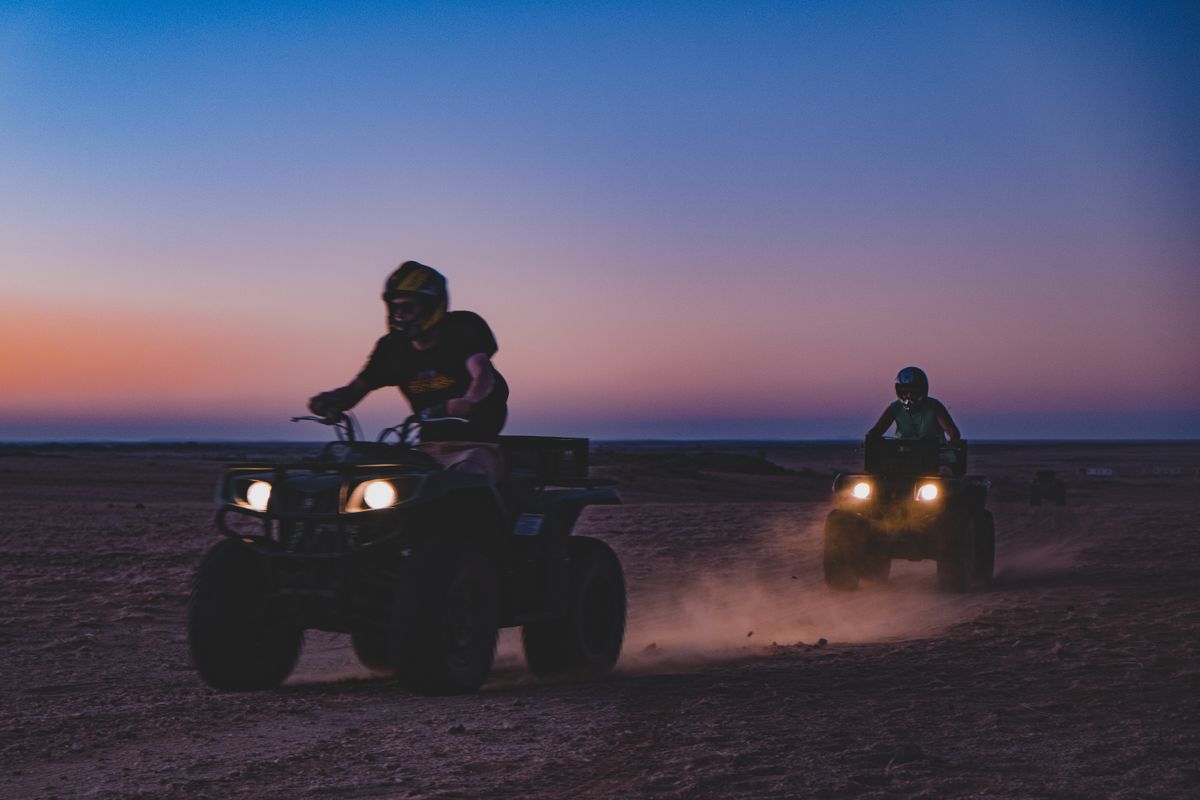 Two riders on quad bikes crossing open desert at dusk, headlights on, dust trailing