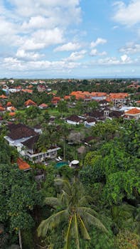 Aerial view of a tropical village with lush greenery and red roofs under a partly cloudy sky.