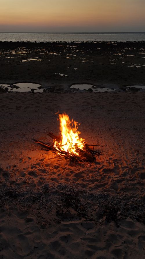 Free Glowing bonfire on a sandy beach during sunset with ocean in the background. Stock Photo