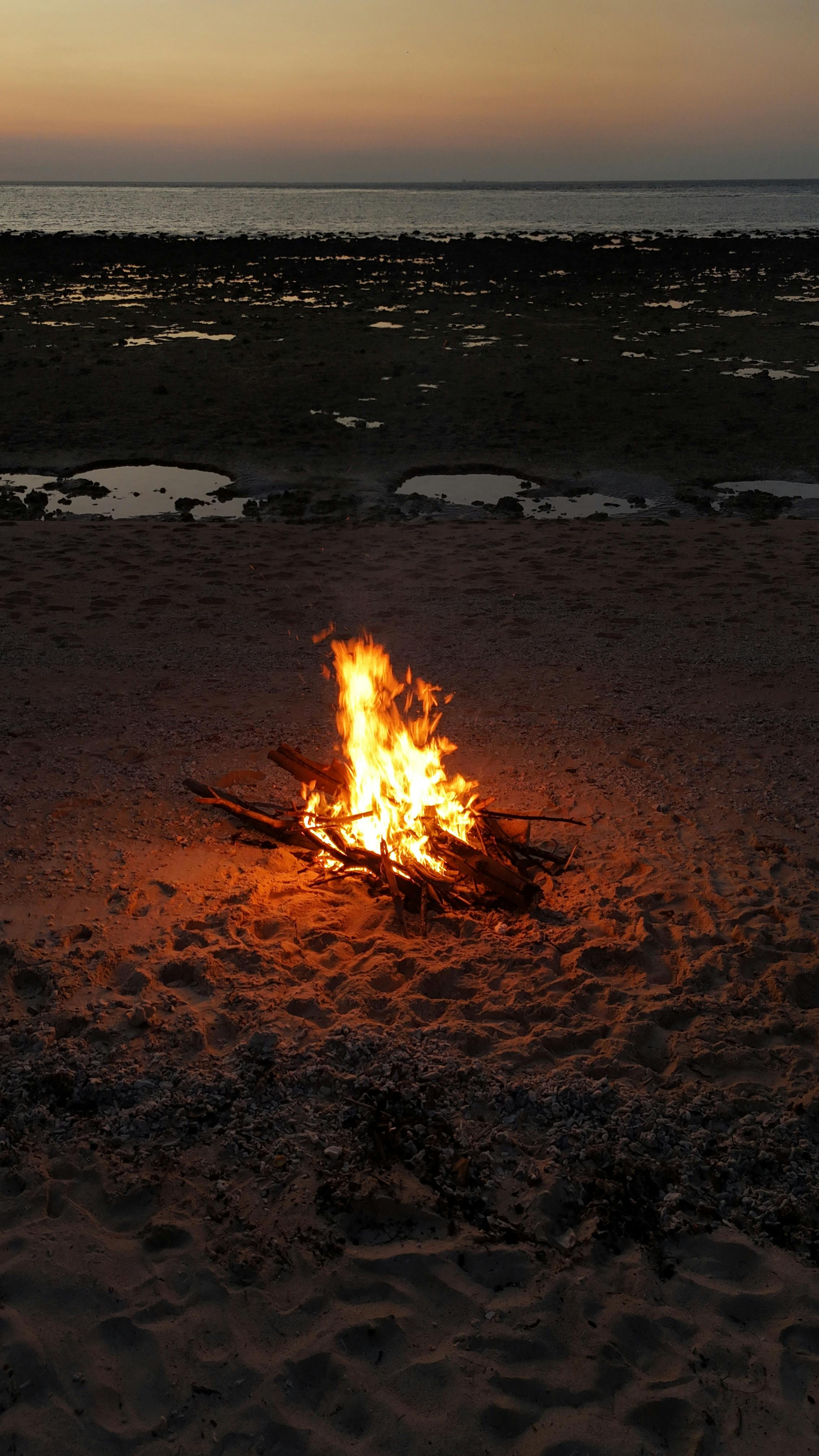 Gratis Falò acceso su una spiaggia sabbiosa al tramonto, con l'oceano sullo sfondo. Foto a disposizione