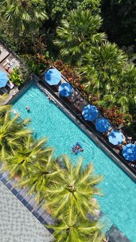 Aerial shot of a vibrant hotel pool with sunbathing guests surrounded by lush greenery and blue umbrellas.