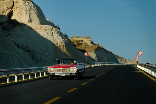 A red vintage convertible car driving on a scenic highway with rocky cliffs under a clear blue sky.