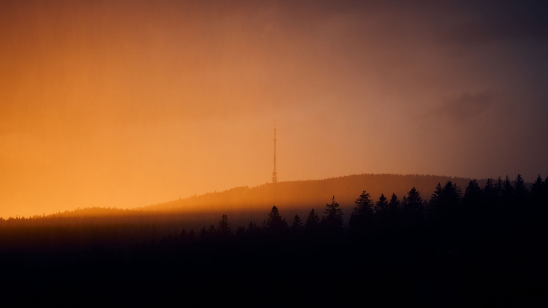 Dramatic sunset over silhouetted forest in Germany with a distant tower under cloudy skies.