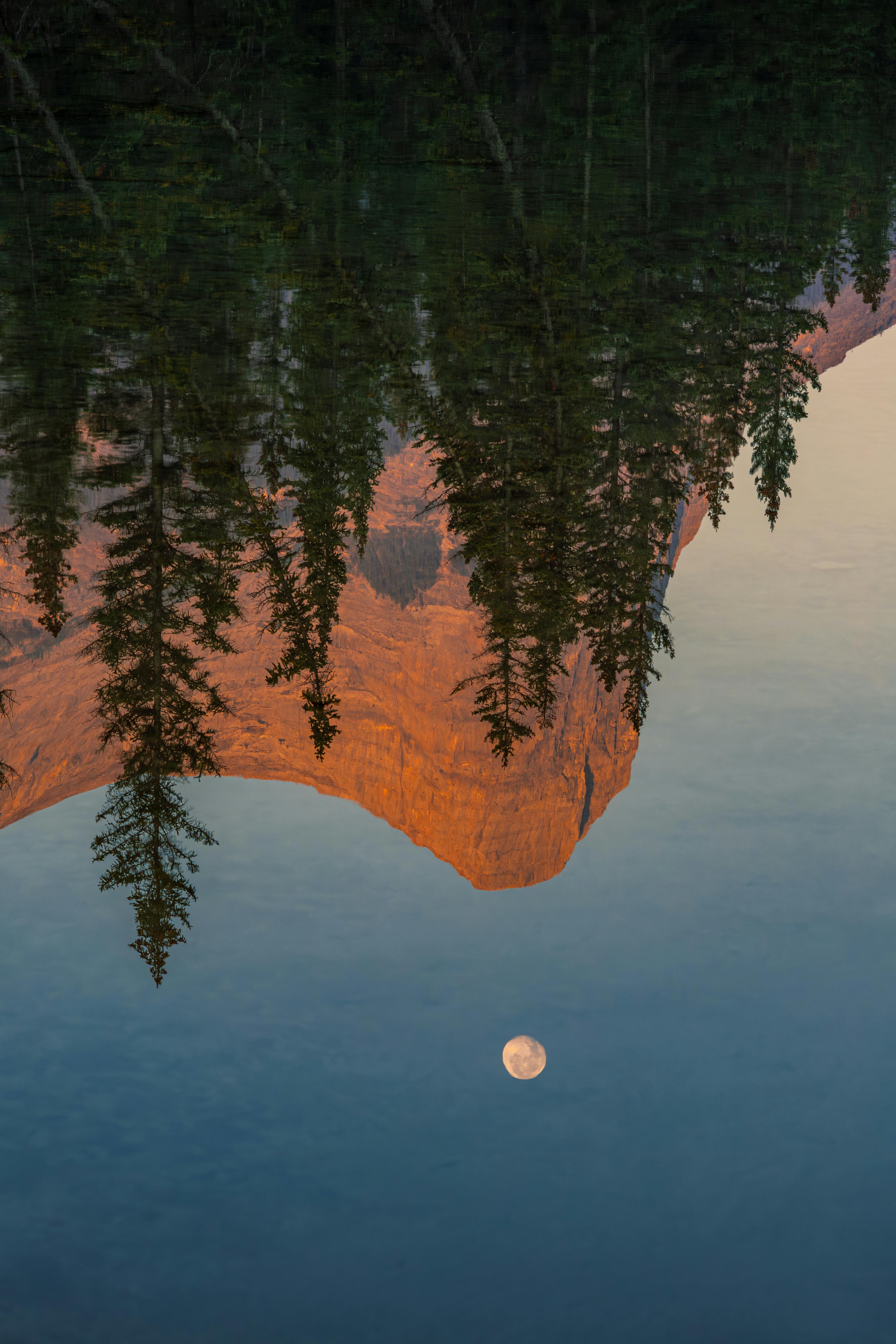 Serene reflection of moon and mountains in calm water at dawn in Canmore, Alberta.