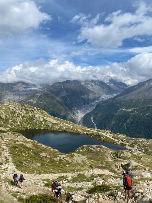 Free Hikers explore a stunning alpine landscape with a serene lake and majestic peaks under a bright blue sky. Stock Photo