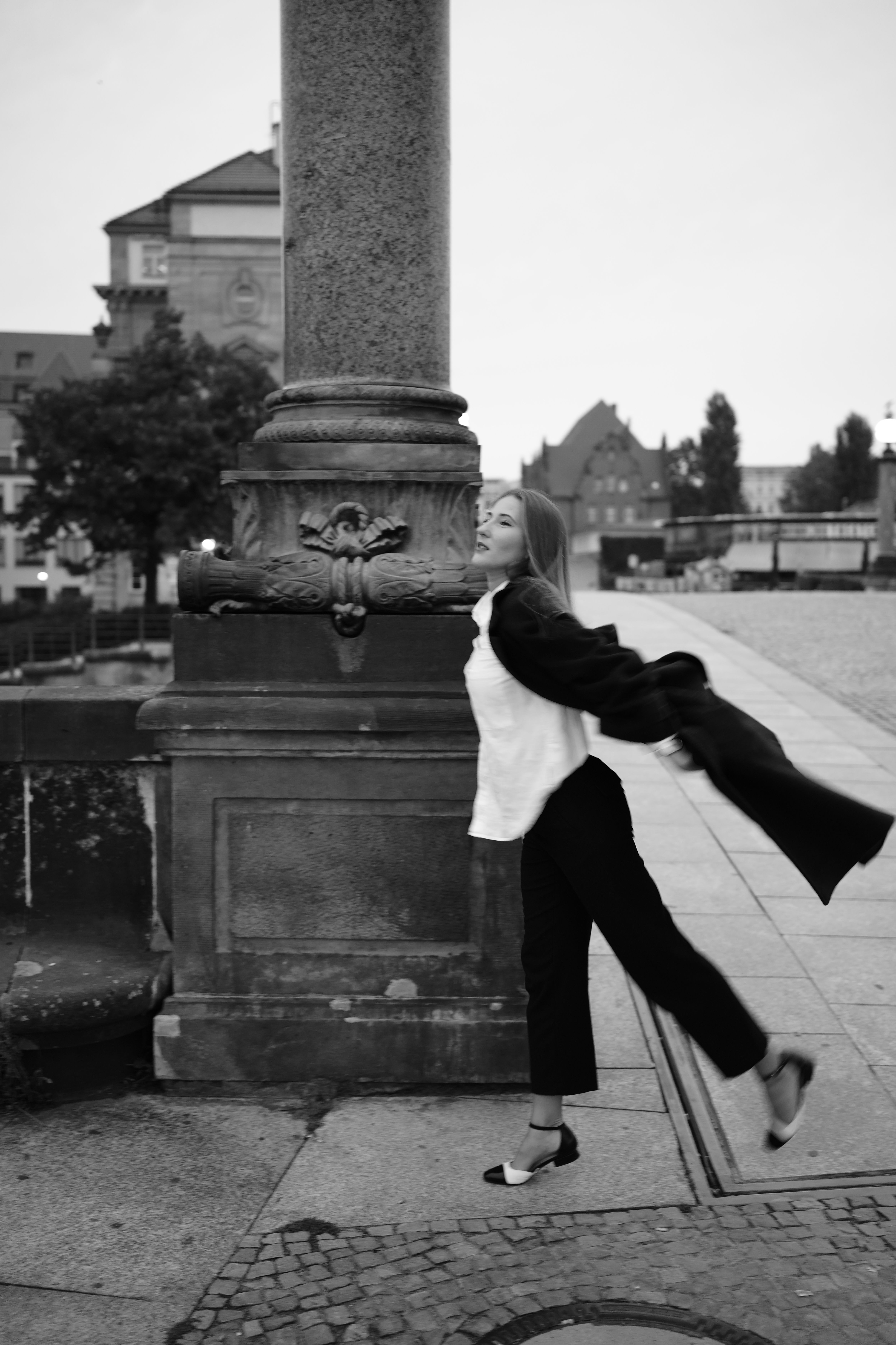 Elegant woman joyfully posing against a pillar in black and white street scene.