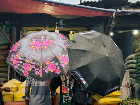 Two people under black and floral umbrellas at an outdoor market on a rainy day.