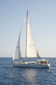 A white sailboat with people on board sailing on a clear, sunny day in open ocean waters.