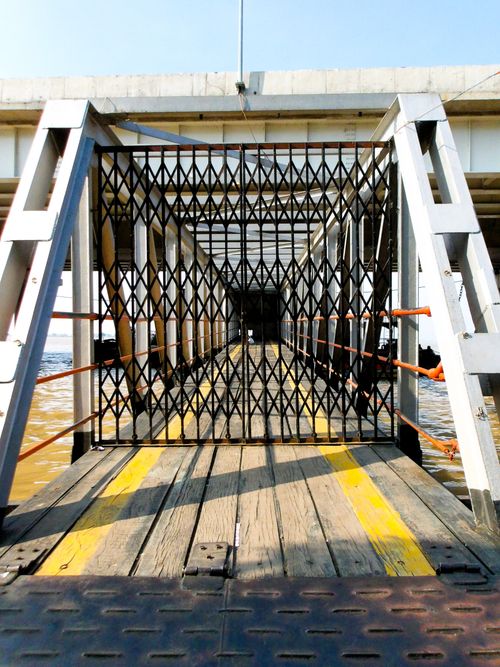 Free A wooden bridge with a metal grate leads over a water body under a bright sky. Stock Photo