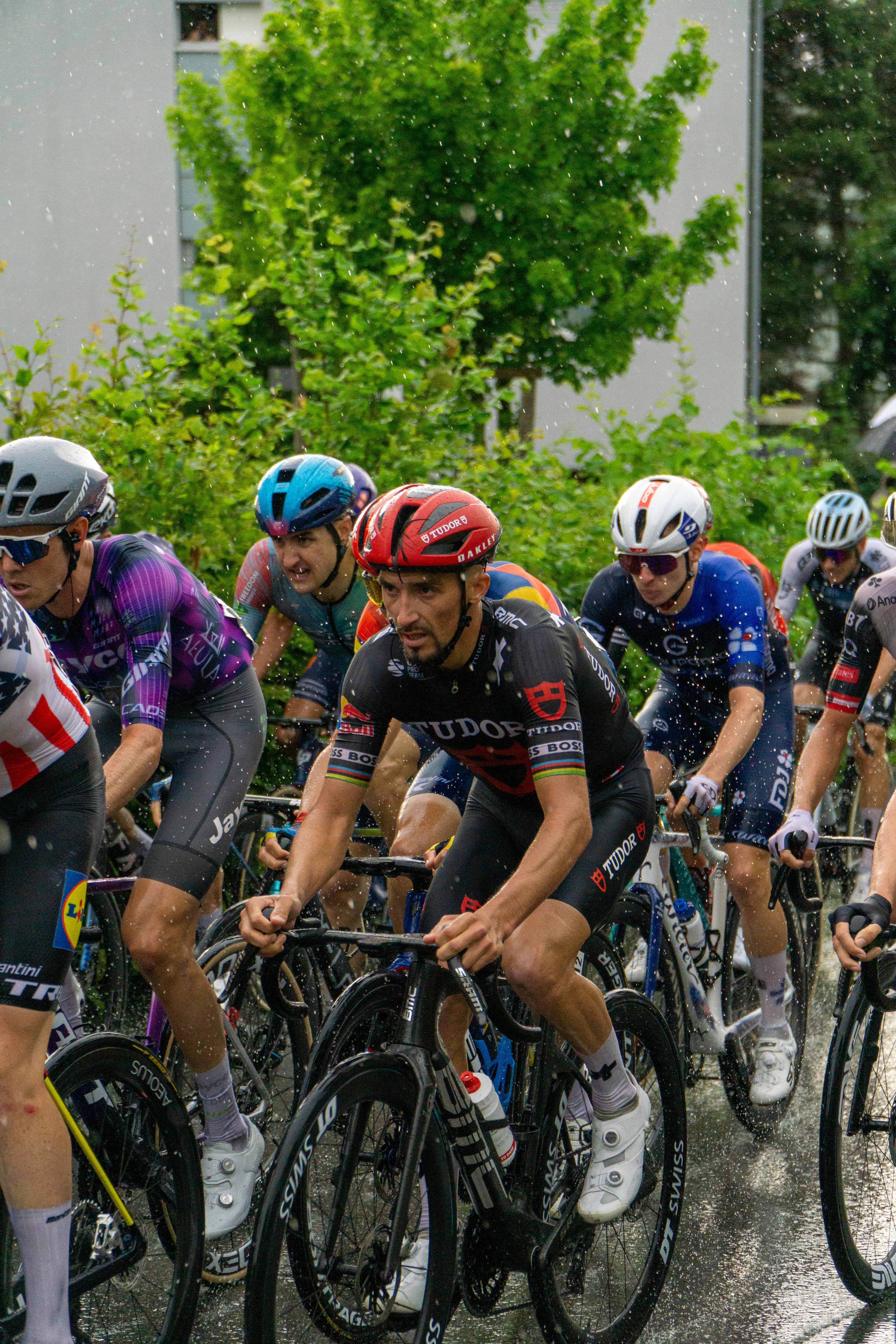 Pelotão de ciclistas profissionais disputando etapa sob chuva intensa em estrada molhada