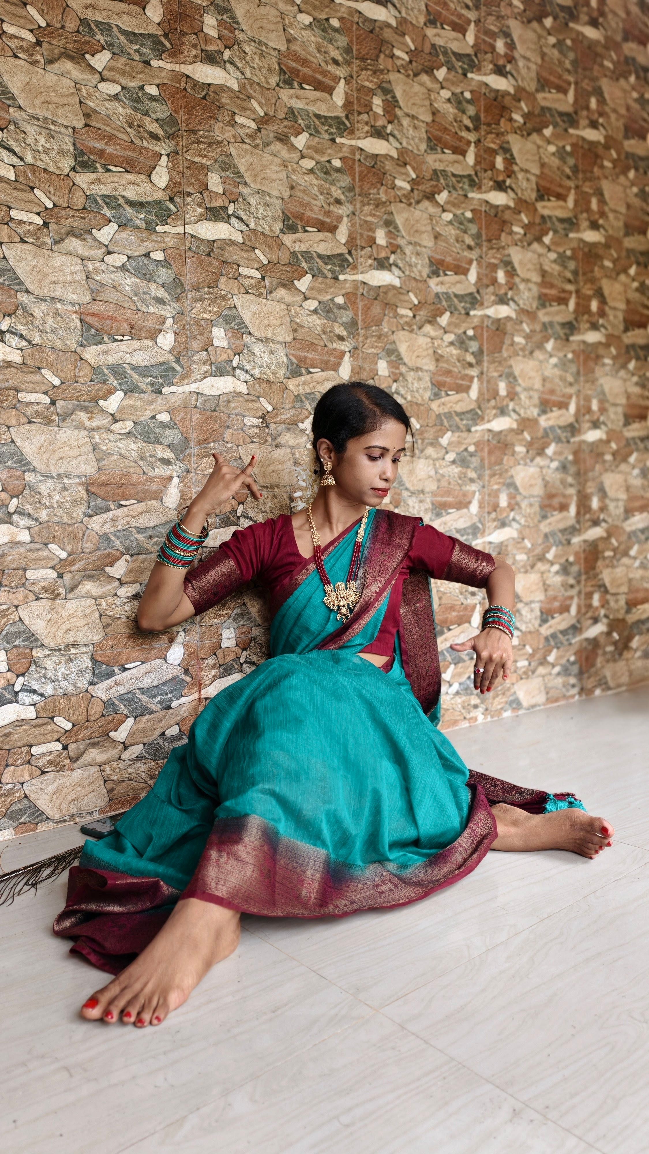 Elegant woman in traditional teal and maroon saree, relaxing indoors against a textured stone wall.