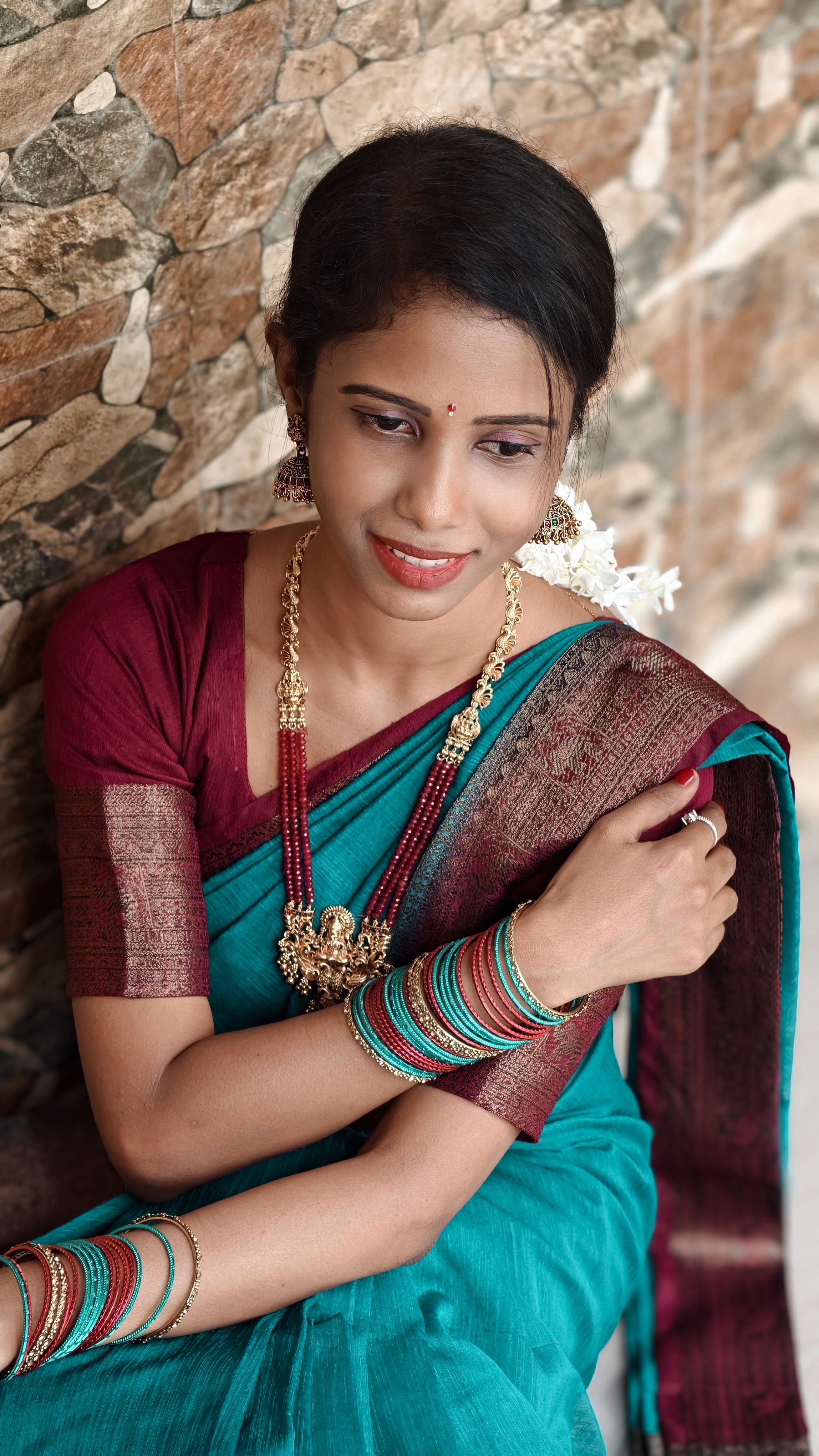 Portrait of a smiling woman in a vibrant saree and gold jewelry, seated against a stone wall.